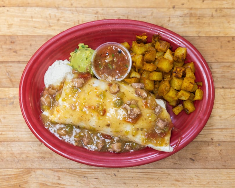 A breakfast burrito smothered in green chili sauce, with a side of crispy potatoes, guacamole, and sour cream.