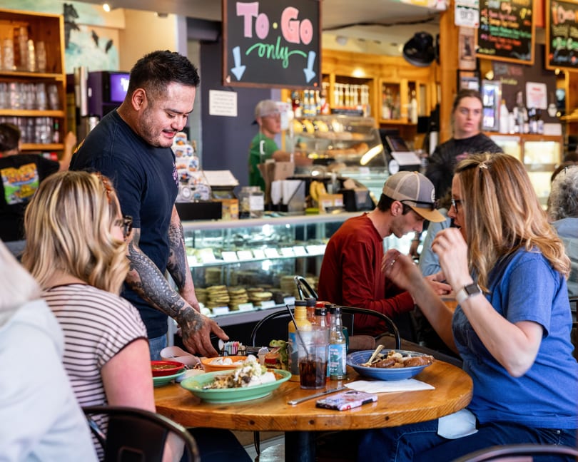 A server with tattoos delivers food to customers seated at a wooden table in a busy restaurant.