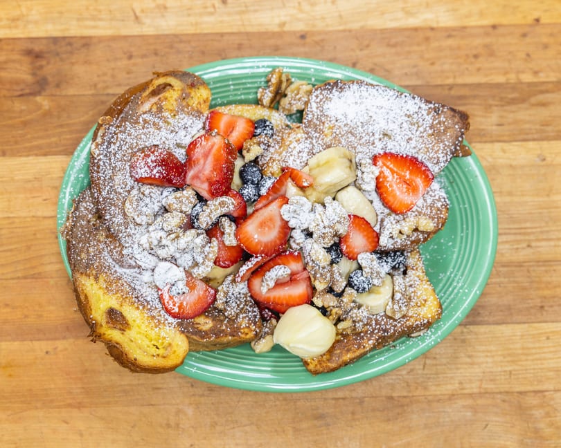 French toast topped with strawberries, blueberries, bananas, walnuts, and powdered sugar on a green plate.