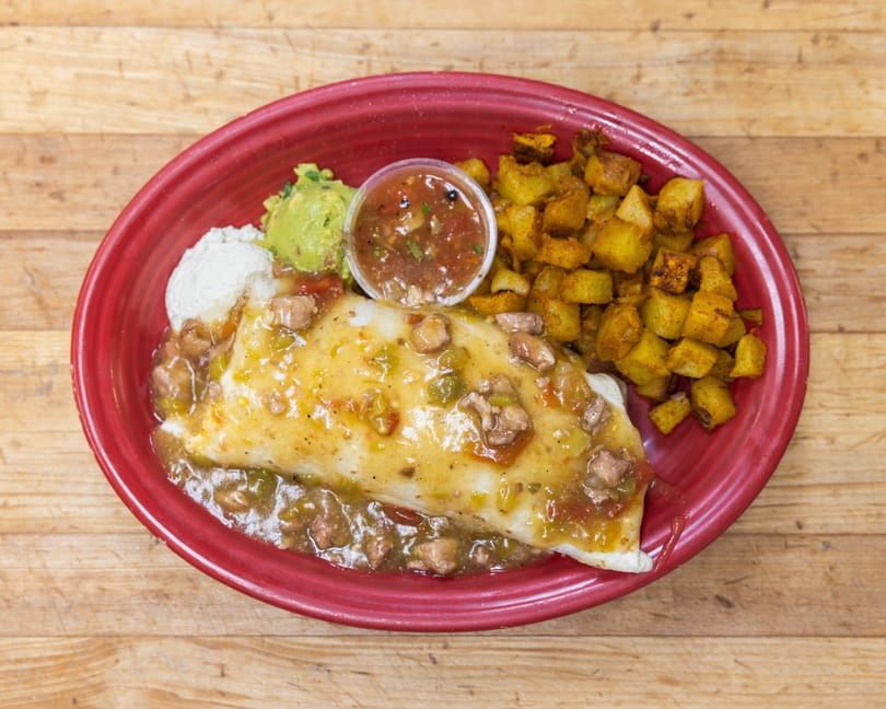 A breakfast burrito smothered in green chile sauce, with a side of potatoes, guacamole, sour cream, and salsa.