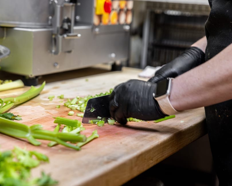 Chef in black gloves chopping celery on a wooden cutting board in a commercial kitchen.