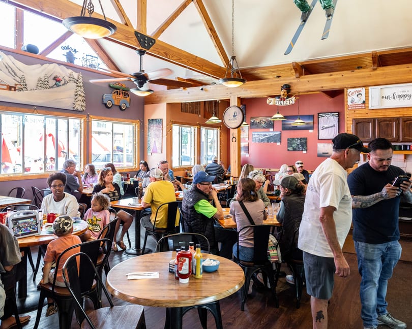 A bustling restaurant interior with many patrons seated at tables, decorated with mountain-themed art and skis.