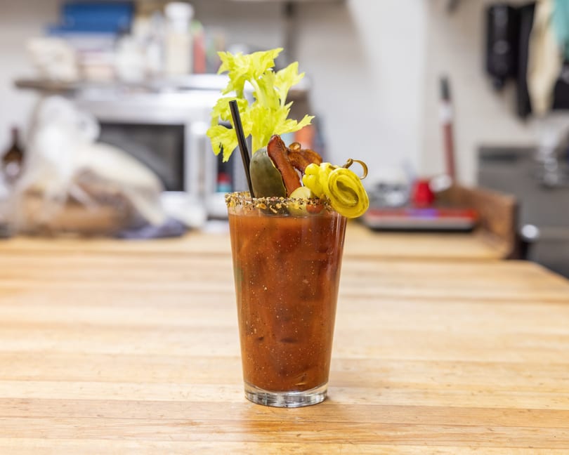 A Bloody Mary cocktail garnished with celery, bacon, pickles, and peppers on a wooden table.