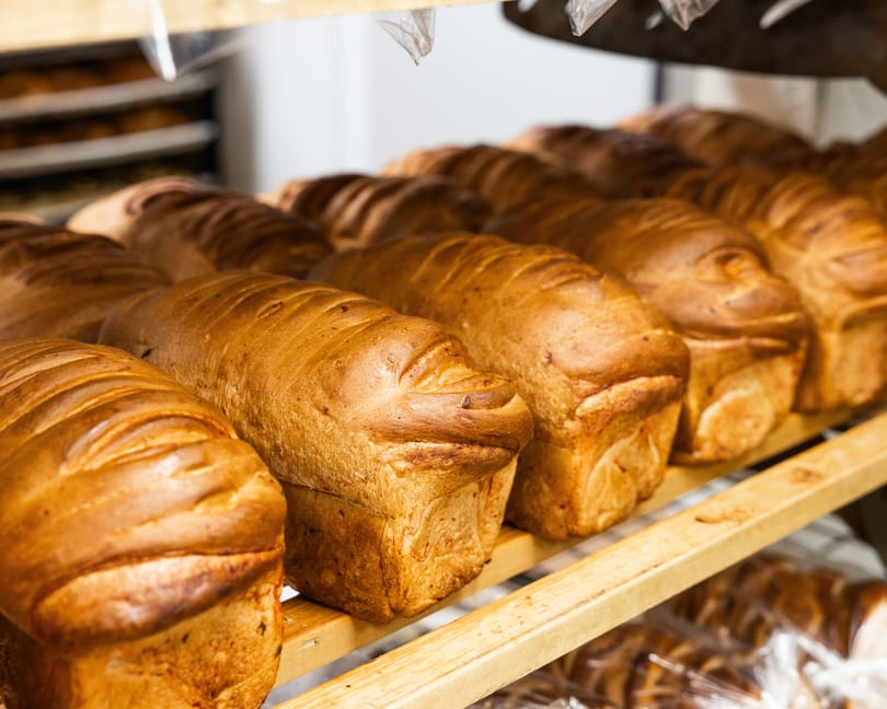 Rows of freshly baked, golden-brown bread loaves sit on wooden shelves in a bakery.