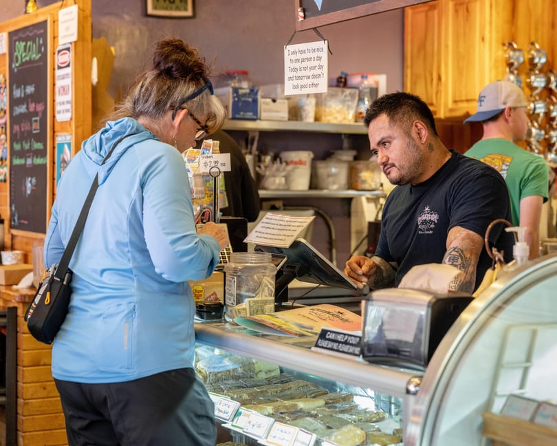 A customer in a blue hoodie orders from a tattooed barista behind a pastry display case.