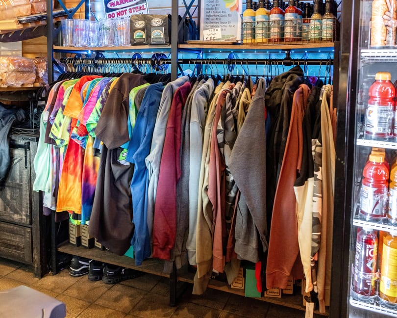 A rack of colorful tie-dye and solid-colored shirts for sale, with drinks and sauces nearby.