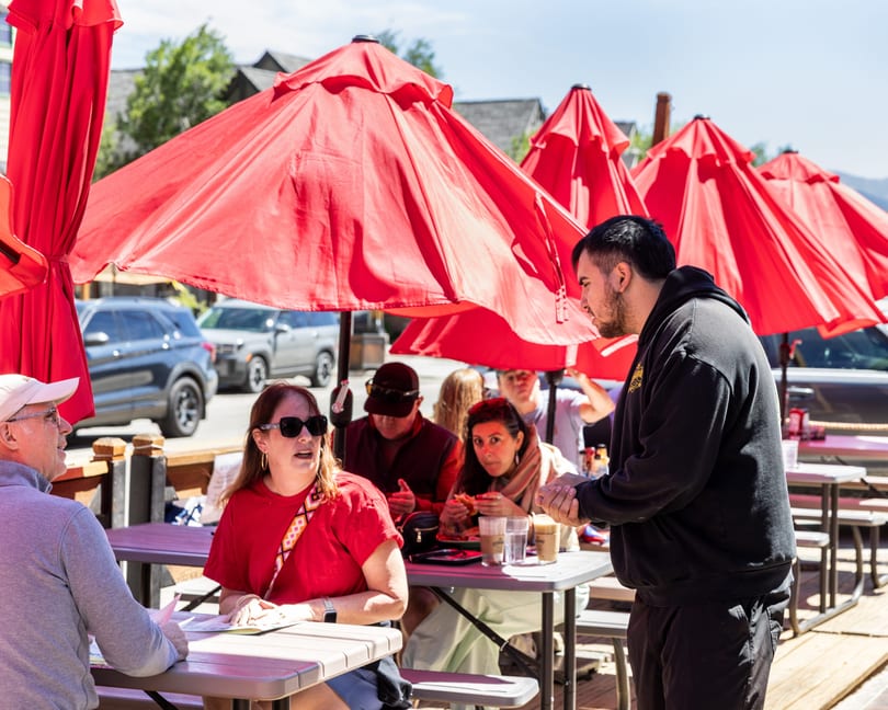 A server talks to customers seated at outdoor tables under red umbrellas on a sunny day.