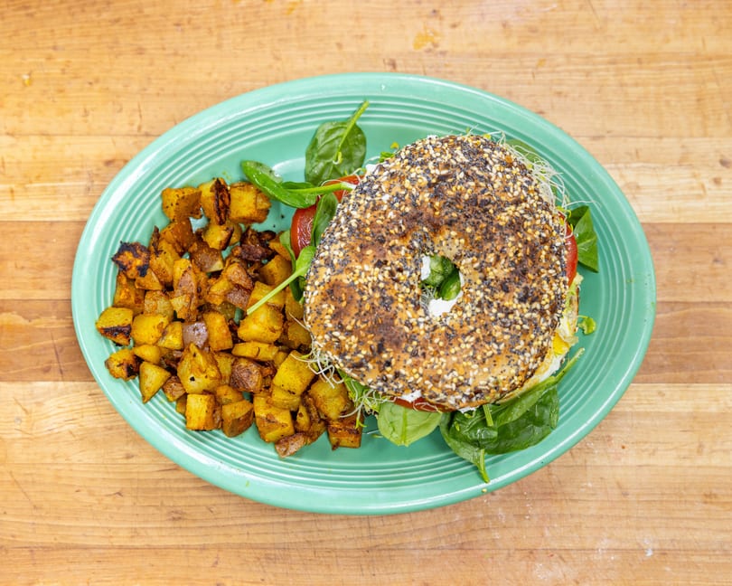 Everything bagel sandwich with egg, spinach, tomato, and sprouts, served with roasted potatoes.