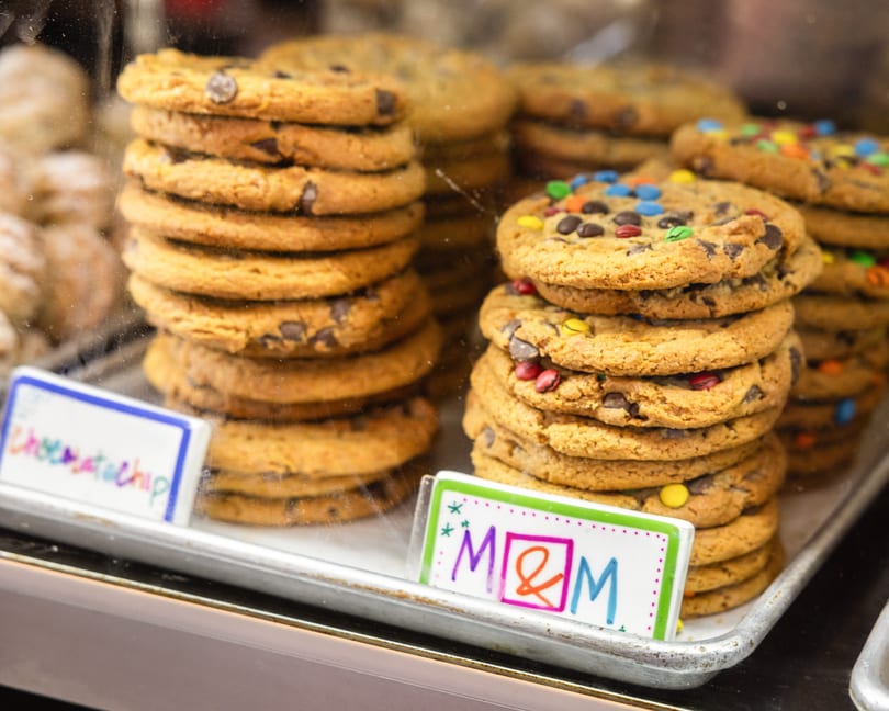 Stacks of chocolate chip and M&M cookies displayed in a glass bakery case.