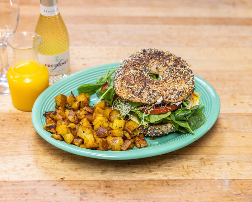 Everything bagel sandwich with spinach, tomato, sprouts, and avocado, served with roasted potatoes and drinks.