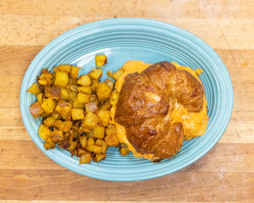 Croissant sandwich with creamy orange filling and a side of crispy breakfast potatoes on a blue plate.